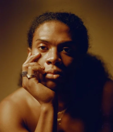 Portrait of a black man resting his chin on his hand with rings on his fingers against a warm-toned background