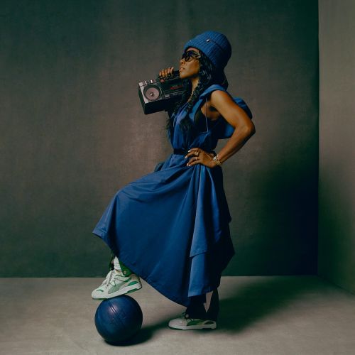 Woman in blue dress and hat balances on a ball, holding boombox in a studio setting, showcasing fashion and music elements.