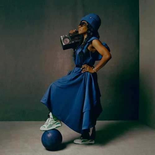 Woman in blue dress and hat balances on a ball, holding boombox in a studio setting, showcasing fashion and music elements.