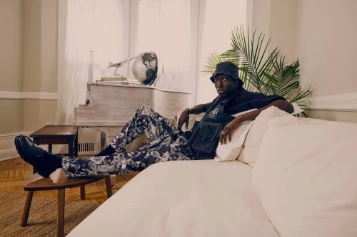 Black man wearing a bucket hat and patterned trousers seated on a white sofa in a light filled living room