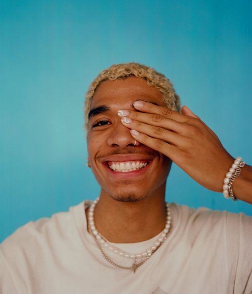 Portrait of a happy young adult with blond hair, pearl necklaces and bracelets, and wearing a white t-shirt against a light blue background.