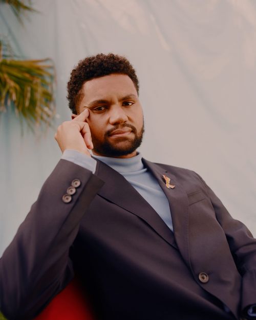 Portrait of an adult man with curly hair wearing a suit, turtleneck, and a lapel pin with a teal and white patterned backdrop.