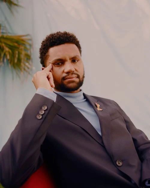 Portrait of an adult man with curly hair wearing a suit, turtleneck, and a lapel pin with a teal and white patterned backdrop.
