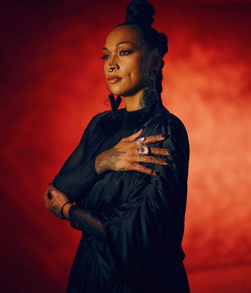 Portrait of an adult female with braided hair, a black dress, and hand tattoos against a red background in a studio setting.