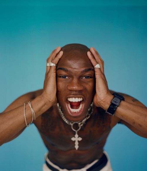 A shirtless, bald African American man with a silver chain and rings is screaming with hands to his head against a teal backdrop.