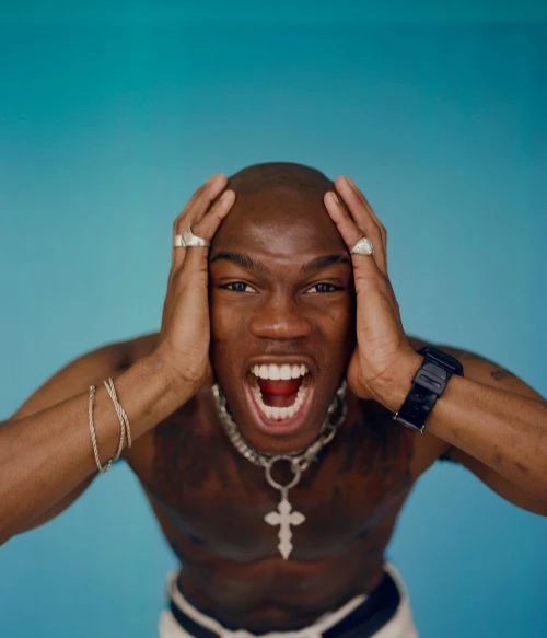 A shirtless, bald African American man with a silver chain and rings is screaming with hands to his head against a teal backdrop.