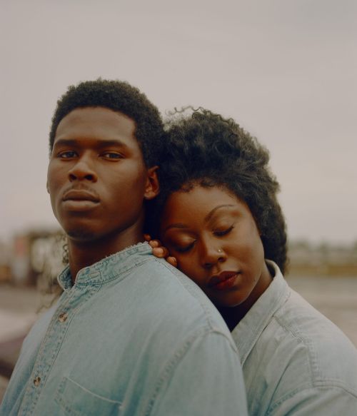 Portrait of a couple wearing matching denim shirts. The woman leans on the man's shoulder against a grey outdoor background.