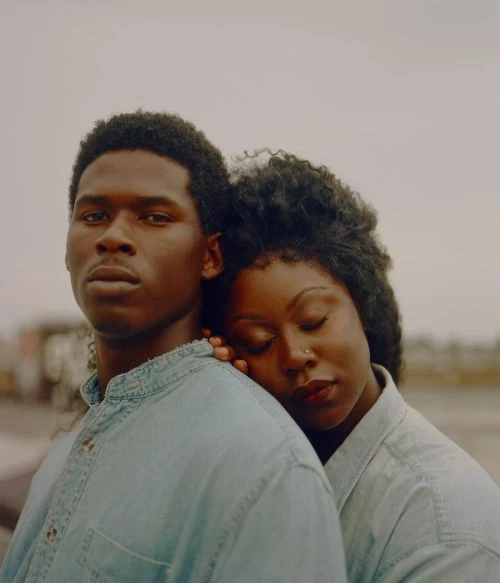 Portrait of a couple wearing matching denim shirts. The woman leans on the man's shoulder against a grey outdoor background.