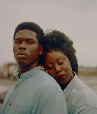 Portrait of a couple wearing matching denim shirts. The woman leans on the man's shoulder against a grey outdoor background.
