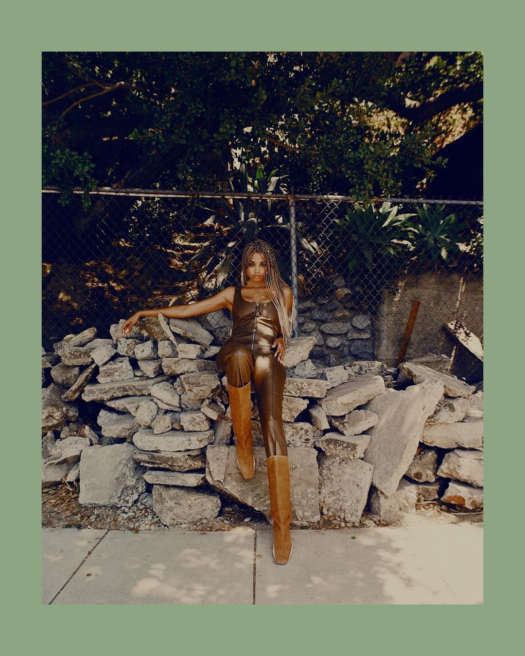 Adult woman poses in a gold jumpsuit and brown boots, leaning against rocks in an outdoor urban setting with green foliage backdrop.