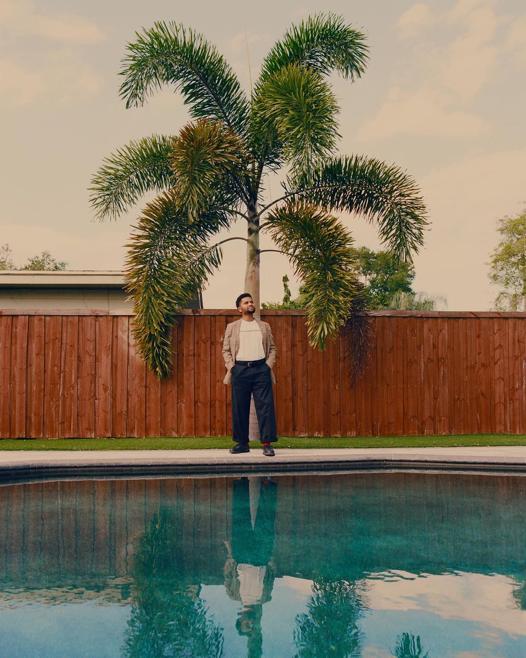 A man standing by a swimming pool in front of a wooden fence and a palm tree, photographed by Kendall Bessent.