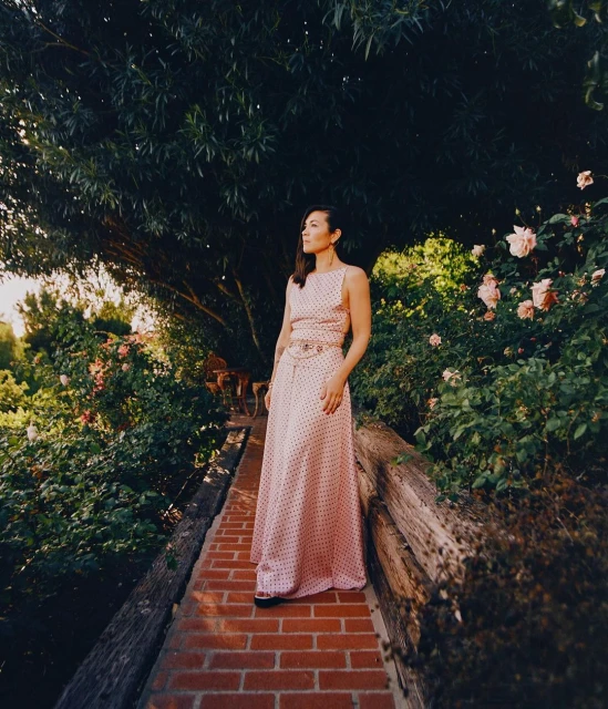 Adult woman in a pink polka dot dress stands on a red brick path in a verdant garden setting with roses and trees in the background.
