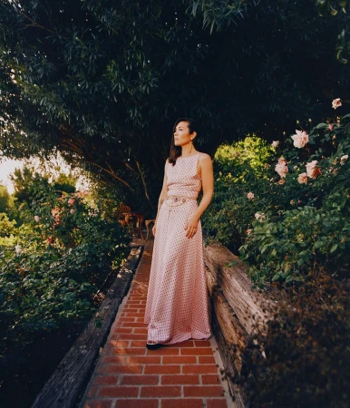 Adult woman in a pink polka dot dress stands on a red brick path in a verdant garden setting with roses and trees in the background.