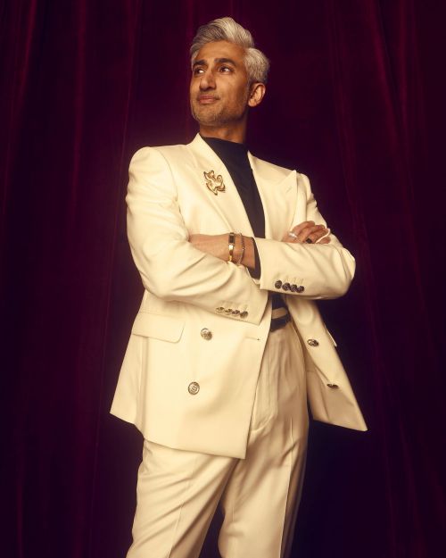 Adult male in a cream-colored suit poses in a studio. He has silver hair and his arms are folded, with a dark-red curtain backdrop.