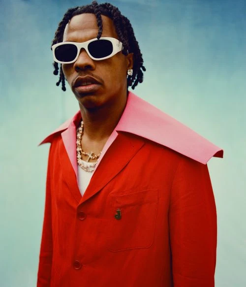 Young man poses in red jacket and sunglasses against gradient blue backdrop. He wears chains, earrings, and has braided hair.