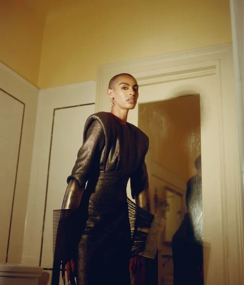 An adult woman with a shaved head wears an elegant dark, sparkly dress with padded shoulders in an indoor hallway setting.