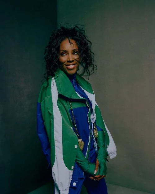 Portrait of an adult woman smiling with curly hair, wearing a blue jumpsuit and color-blocked green and blue jacket indoors.