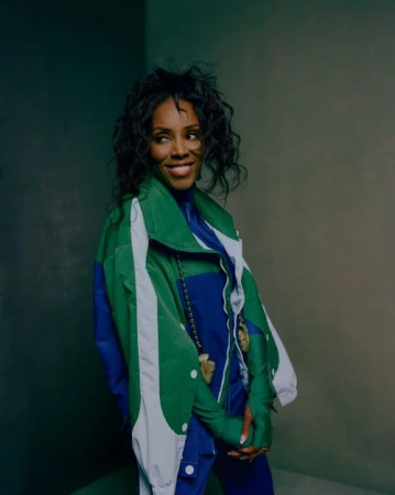 Portrait of an adult woman smiling with curly hair, wearing a blue jumpsuit and color-blocked green and blue jacket indoors.