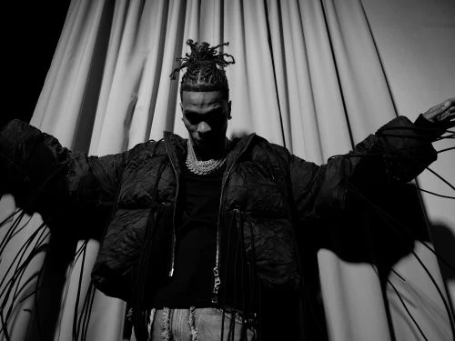 Black man wearing a dark puffer jacket with outstretched arms standing in front of draped fabric