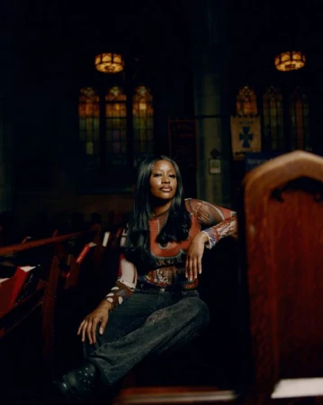Stylish woman with long black hair poses seated in church pew with stained-glass windows in background under dim ambient light.