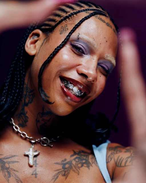 Close-up of a smiling adult female with box braids, eyebrow piercing, lip gloss, and a silver cross necklace against a purple background.