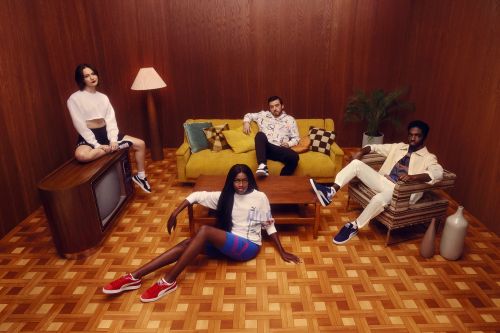 Four people posing in an indoor studio setting with vintage decor, including a sofa, wood paneled walls, and retro television set.