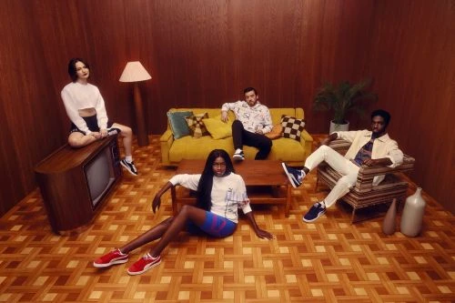Four people posing in an indoor studio setting with vintage decor, including a sofa, wood paneled walls, and retro television set.