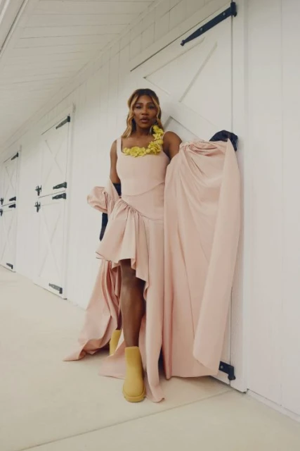 Serena Williams poses in a light pink dress with yellow floral accents, paired with tan boots, against a white paneled barn wall.
