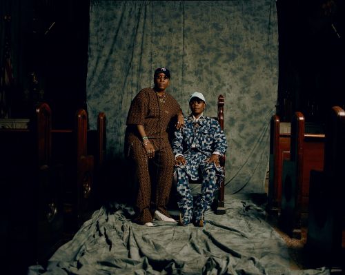 Two adult individuals are seated indoors on a draped surface with a mottled backdrop, both in patterned outfits and hats, facing forward.