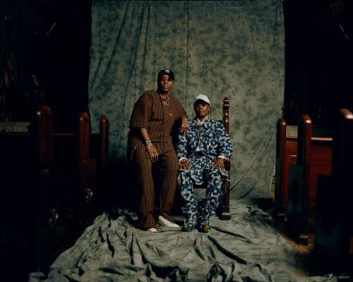 Two adult individuals are seated indoors on a draped surface with a mottled backdrop, both in patterned outfits and hats, facing forward.