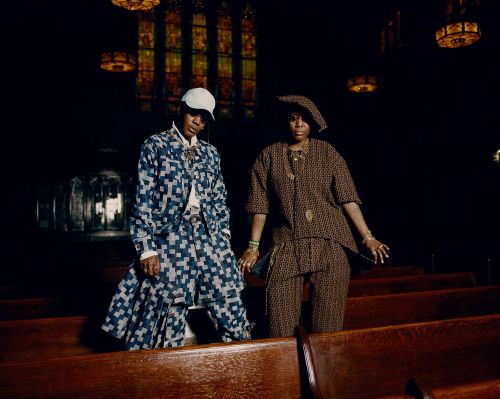 Two women in patterned outfits stand in a church. One wears blue, the other brown. Stained-glass windows and wooden pews form the background.