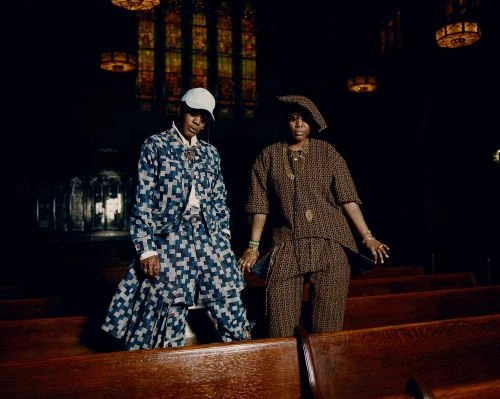 Two women in patterned outfits stand in a church. One wears blue, the other brown. Stained-glass windows and wooden pews form the background.