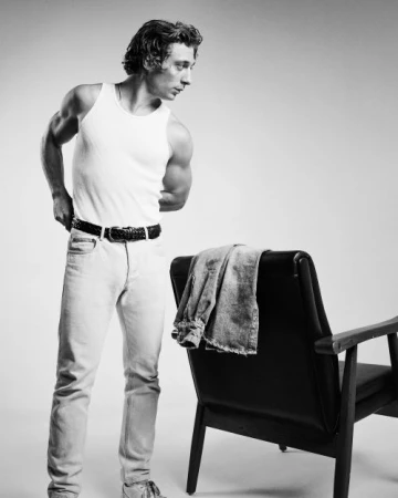 Man wearing a white tank top and light jeans standing next to a black leather chair in a studio