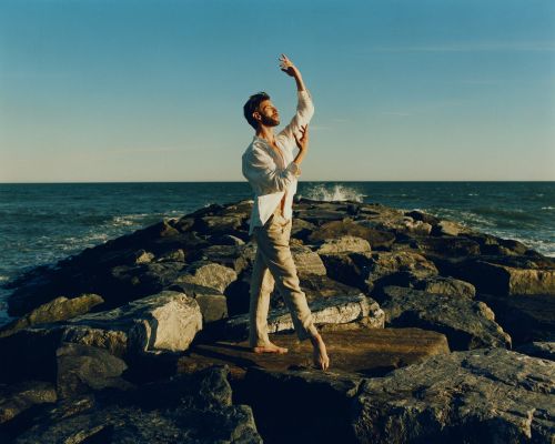 A lone male dancer in a white shirt and tan pants gracefully poses barefoot on a rocky jetty by the ocean under a clear blue sky.