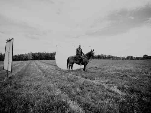 Monochrome shot of an adult riding a horse in a grassy field, with lighting equipment and trees visible in the background.