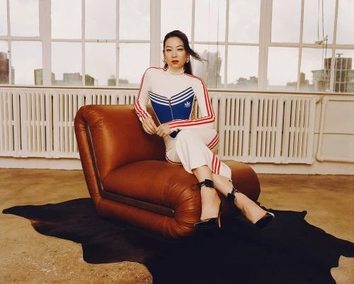 Stylish woman in a white Adidas tracksuit sits on a brown leather chair in front of a large window in a minimalist apartment.