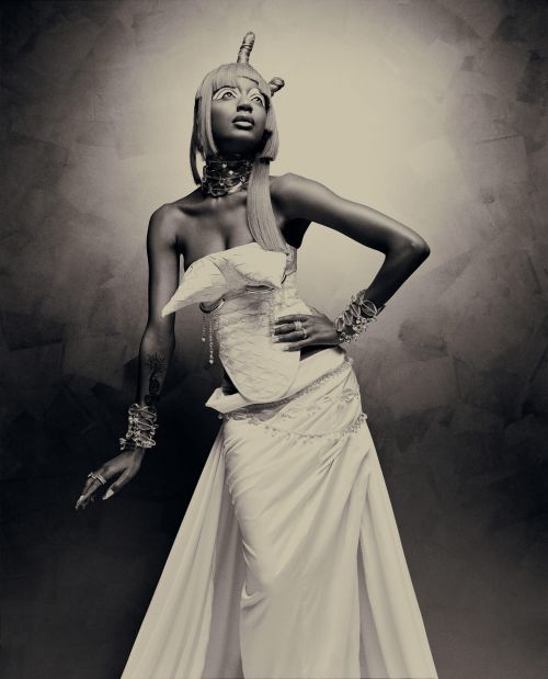 Elegant portrait of a woman in an elaborate white gown and headpiece, striking a pose against a textured, neutral-toned backdrop in the studio.