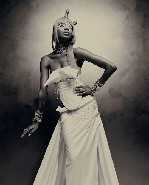 Elegant portrait of a woman in an elaborate white gown and headpiece, striking a pose against a textured, neutral-toned backdrop in the studio.