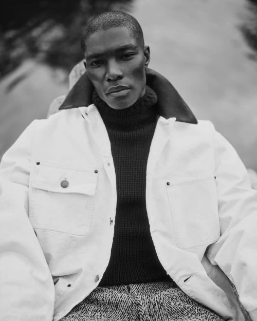 Monochromatic portrait of a Black male model in a white work jacket and dark turtleneck sweater, posing outdoors.