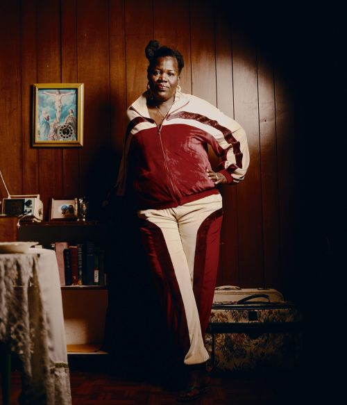 Portrait of a Black woman in a maroon-and-cream tracksuit standing indoors. The scene is illuminated by a bright overhead light.