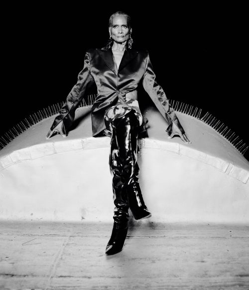 Elegant older woman poses in a satin blazer and patent leather boots, seated against a dark background, studio lighting.