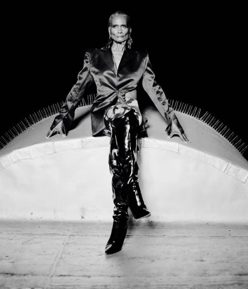 Elegant older woman poses in a satin blazer and patent leather boots, seated against a dark background, studio lighting. 