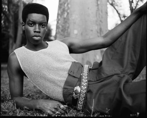 Monochrome portrait of a Black adult model lounging outdoors in a sleeveless top and textured pants with a decorative belt.