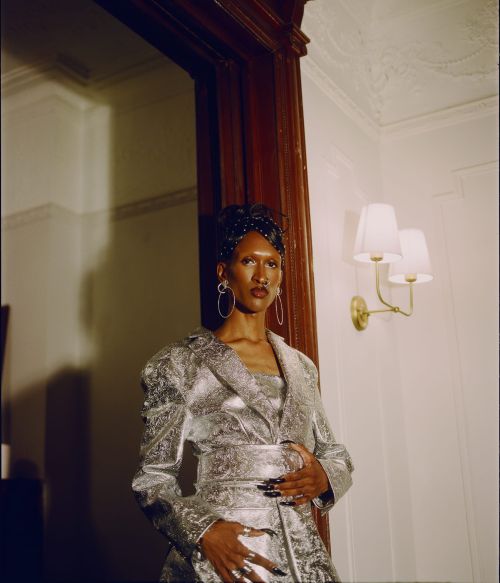 Adult female poses indoors in silver metallic dress and jacket with hoop earrings, decorative hair clips, and dark lipstick and nail polish.