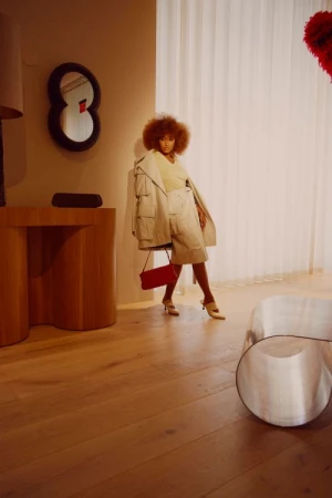 Black woman with afro hair in beige jacket and shorts holding a red handbag indoors by Kendall Bessent