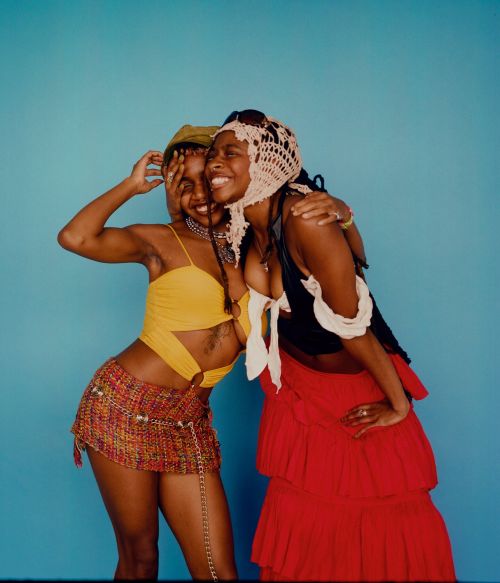 Two smiling Black women pose in front of a light blue backdrop. One wears a yellow top and multicoloured skirt; the other wears a red skirt.