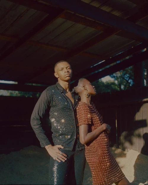 A stylish couple poses in an indoor-outdoor setting; the Black man wears a sequined shirt, and the Black woman wears a sequined dress.