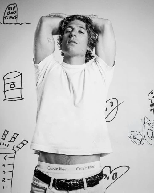 Black and white studio shot of a man with curly hair posing in underwear and a t-shirt, against a wall with cartoon drawings.
