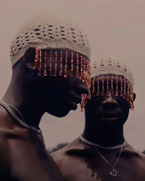 Two dark-skinned male adults wear beaded caps and silver necklaces in a portrait with muted outdoor lighting, creating an artistic, cultural image.