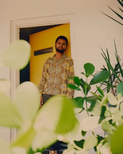 Stylish man in patterned shirt stands near yellow door framed by indoor plants, bathed in soft light, creating a vibrant, relaxed scene.